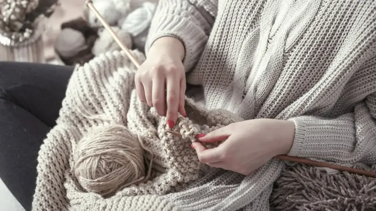 Ręce dziergają ciepły sweter, a obok leżą kłębek wełny i drewniane druty. W tle widać więcej włóczki, jakby przygotowywano kolejne kokonki.