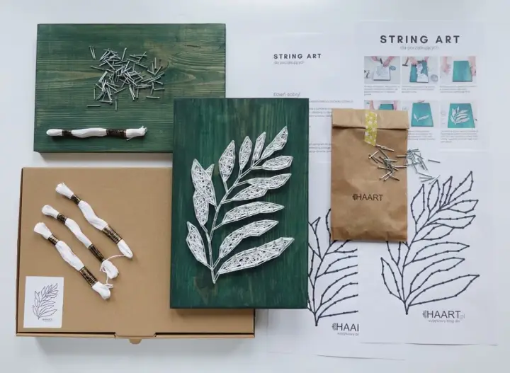 Zestaw do tworzenia obrazów string art z motywem liścia. W pudełku deska, gwoździe, biała nić i instrukcja.